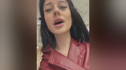 Media: Video of a young woman with long black hair, wearing a red leather jacket, with a sultry expression and slightly parted lips, indoors with a blurred background.