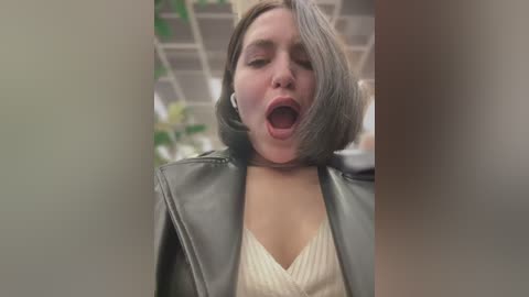 Media: Video of a young woman with light skin, short gray hair, and a black leather jacket, mouth open in apparent surprise or excitement. Background shows a blurry office setting with plants and white ceiling tiles.