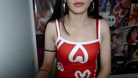 Media: Video of a young woman with pale skin and long black hair, wearing a tight red tank top with white McDonald's logo, silver hoop earrings, and a black armband, standing in a room with colorful posters in the background.