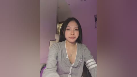 Media: A video of an Asian woman with long black hair, wearing a gray cardigan with a low neckline, seated in a room with purple walls.