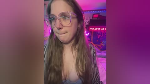 Media: Video of a light-skinned woman with long brown hair, wearing large glasses and a striped shirt, smiling slightly. Background features a colorful, neon-lit room with gaming equipment and posters.