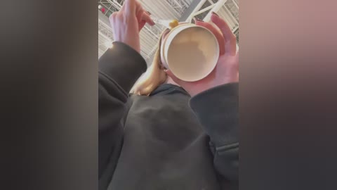 Media: Video of a person wearing a black hoodie, holding a white coffee mug with a yellow and brown bunny design, in an industrial setting with exposed beams and pipes.