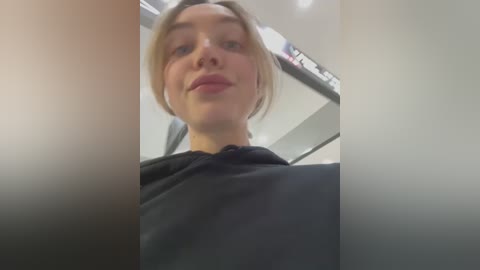 Video of a woman with fair skin, blonde hair, and light makeup, wearing a black top, taken from a low angle, with blurred background.