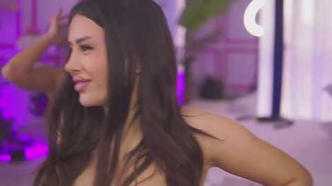 Media: Video of a topless woman with long, dark hair, light skin, and a slender physique, posing indoors. The background features blurred purple lighting and green foliage, suggesting a modern, softly lit setting.