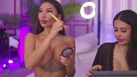 Media: Video of two topless women in a studio; one applying makeup with a yellow stick, the other holding a compact mirror, both with fair skin and dark hair.