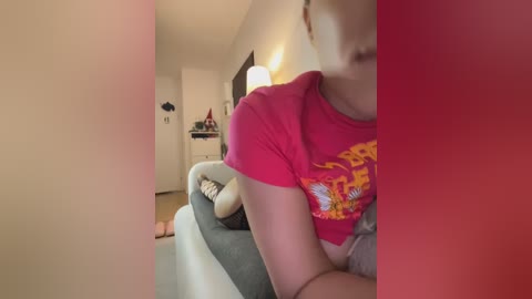 Media: Video of a woman in a red t-shirt, sitting on a gray couch, with a blurry background showing a white door, lamp, and a cat.