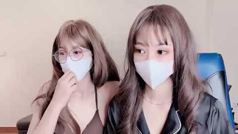 Media: Video of two Asian women with long brown hair, wearing face masks and glasses, sitting side-by-side in a room with beige walls.