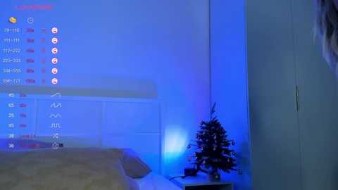 Media: Video of a dimly lit bedroom with blue ambient lighting, featuring a bed with beige bedding, a small Christmas tree, and a wall calendar.