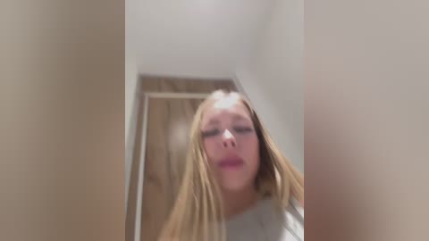 A video of a blonde woman with long hair and a neutral expression, captured from a low angle, standing in a hallway with light brown walls and a wooden door.