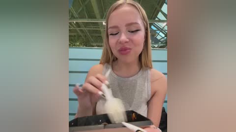 Video of a young blonde woman with fair skin, wearing a gray tank top, puckering her lips, applying white powder with a brush to her face.