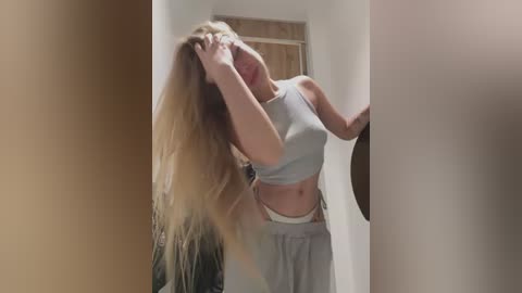 Video of a blonde woman with long hair, wearing a gray crop top and matching loose pants, standing in a doorway, adjusting her hair.