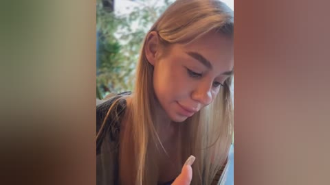 Media: Video of a young woman with long blonde hair, wearing a black top, intently looking at her phone, surrounded by blurred greenery.