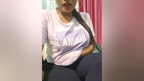 Media: Video of a light-skinned woman with long, straight black hair, wearing a white T-shirt with \"Glamour\" printed in bold letters, sitting in a chair, surrounded by pink and white curtains.