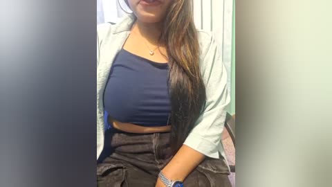 Media: Video of a young woman with long brown hair, wearing a light blue jacket over a navy blue crop top, high-waisted dark denim shorts, and a silver bracelet. She sits in a room with vertical blinds and a light green wall.