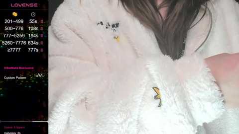 Media: A video shows a young woman with long brown hair, wearing a white fluffy robe with a yellow bunny plushie on the chest. She's indoors, possibly in a bedroom, with a smartphone screen in the background displaying her \"LOUNGE\" status.