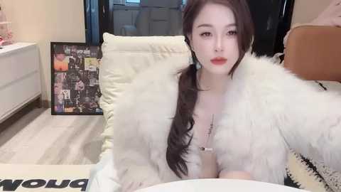 Media: Video of an Asian woman with long black hair in a white fur coat, seated in a modern living room with a framed poster and a brown leather chair.