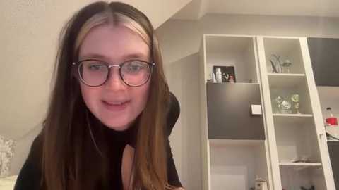 Media: Video of a young woman with long, straight brown hair and glasses, smiling, standing in a modern, light-colored bedroom with built-in white shelves and decorative items.