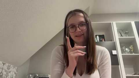 Media: Video of a young Caucasian woman with straight brown hair and glasses, wearing a white ribbed sweater, smiling and making a peace sign with her right hand. She stands in a modern, minimalistic room with a white wall and a grey cabinet.