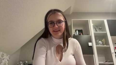 Media: Video of a smiling Caucasian woman with long brown hair, wearing glasses and a white ribbed sweater with a keyhole cutout, standing in a modern, minimalist kitchen with white cabinets and a black fridge.