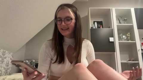 Media: Video of a smiling, fair-skinned woman with long brown hair, wearing glasses, a white top, and holding a phone, in a modern kitchen with white cabinets, gray backsplash, and metallic appliances.