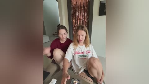 Media: Video of two women, one with shoulder-length blonde hair in a white t-shirt, the other with dark hair in a burgundy t-shirt, crouched on a wooden floor, playing a video game.