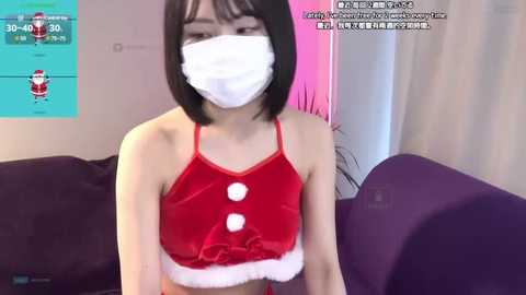 Media: A video of an East Asian woman with short black hair, wearing a red, fuzzy Santa-themed crop top and a white face mask, sitting on a purple sofa, with a live streaming overlay in the background.