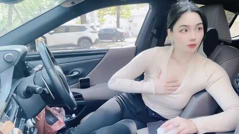 Media: Video of a young Asian woman with fair skin, wearing a beige long-sleeve top and black pants, sitting in a car with a worried expression, holding her chest, car interior visible.