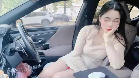 Media: Video of a young Asian woman with long black hair, wearing a beige dress, sitting in a car, yawning, with a blurred parking lot in the background.