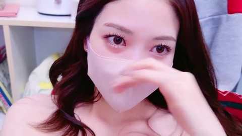 Media: Video of a young Asian woman with long, dark hair, wearing a clear face mask, sitting indoors with a white shelf in the background, holding a finger to her lips.