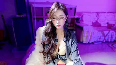 Media: Video of an Asian woman with long brown hair, wearing glasses and a grey jacket over a beige bra, seated on a white fur rug in a dimly lit living room with purple lighting.