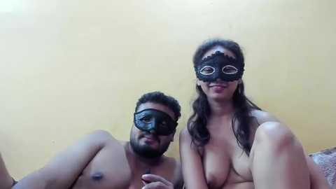 Media: Video of a shirtless Black man with a mustache and a black mask, sitting next to a topless woman with large breasts and a black mask, both with light skin tones, against a beige wall.