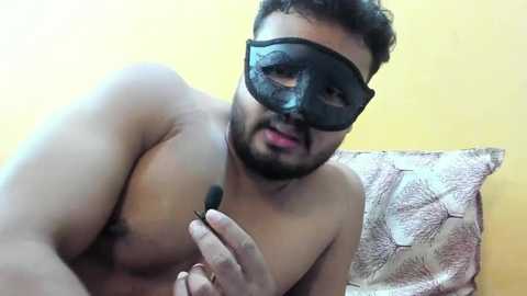 Media: Video of a shirtless South Asian man with a black mask, holding a makeup brush, leaning on a bed with floral pillows against a yellow wall.