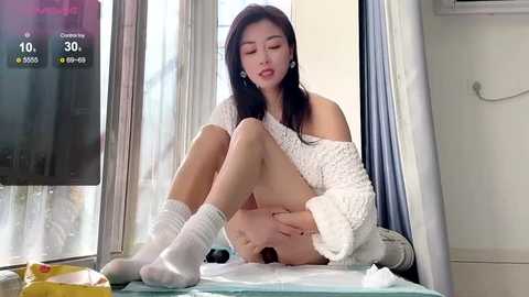 Media: Video of an East Asian woman with long black hair, sitting on a bed, wearing a white off-shoulder sweater, white socks, and no pants. The room has a TV and white walls, with natural light streaming in.