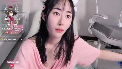 Media: Video of an Asian woman with long black hair, wearing a pink shirt, seated on a white chair in a clinical setting. A digital screen with a game overlay is visible in the background.