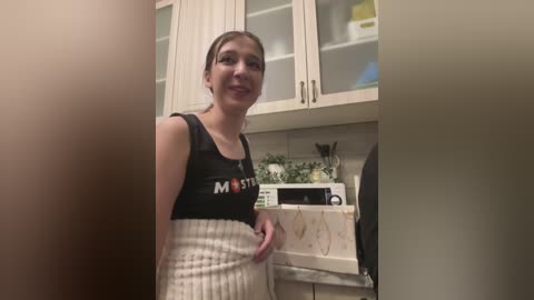 Media: Video of a young woman with light skin, brown hair tied back, wearing a black tank top with \"MOTIVATION\" and a high-waisted white skirt in a modern kitchen with white cabinets, a microwave, and a marble countertop.