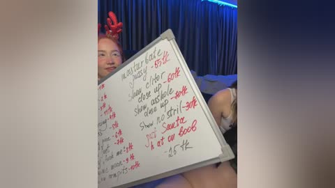 Media: Video of a smiling, fair-skinned woman with red antlers, holding a whiteboard with handwritten text in red and black. She's sitting on a bed with grey curtains in the background, illuminated by blue LED lights.