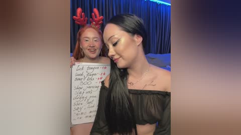 Media: Video of two women, one with red hair and antlers, holding a handwritten sign, the other with long black hair, wearing a black off-shoulder top. Background includes curtains and blue lighting.