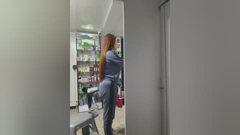 Media: Video of a woman in a blue jumpsuit, standing in a pharmacy, holding a red bag, facing a mirrored wall. Shelves behind her hold various medications and products.