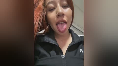 Media: A video of an Asian woman with long, straight red hair and fair skin, sticking her tongue out, wearing a black zip-up jacket with a white logo. The background is blurred, suggesting an indoor setting.