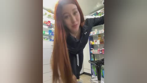 Media: Video of a young woman with long, reddish-brown hair, wearing a black hoodie, leaning over a pharmacy shelf filled with various products.