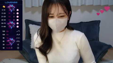 Media: A video of an Asian woman with long, dark hair and a white face mask, wearing a white ribbed sweater, sitting on a blue sofa with a light background.