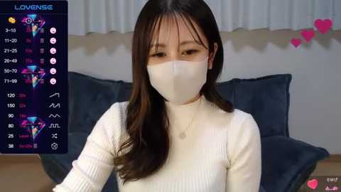 Media: Video of an East Asian woman with long black hair, wearing a white face mask, white turtleneck, and sitting in a dark room with a blue couch.