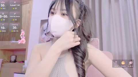 Media: A video of an East Asian woman with long black hair in braids, wearing a white face mask, and a gray knitted top, indoors with a wooden shelf and pink flowers in the background.