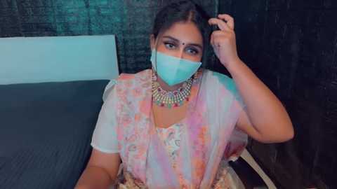 Media: Video of a South Asian woman in a pastel-colored sari, with a turquoise face mask, ornate jewelry, and a hand in her hair, seated on a bed against a dark wall.