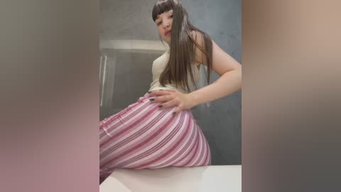 Media: Video of an Asian woman with long black hair, wearing a tight, sleeveless top and striped pink and white pants, seated on a white counter.