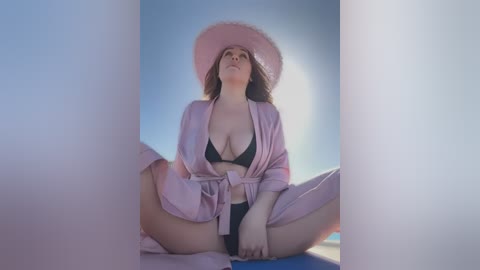 Media: Video of a plus-size woman with fair skin, long brown hair, and a large bust, wearing a pink robe and black bikini, seated with legs spread, against a soft-focus, blue sky background.