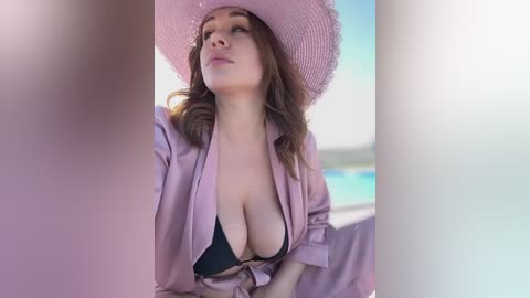 Media: Video of a light-skinned woman with wavy brown hair, wearing a pink hat and open light pink robe, revealing a black bikini top, gazing upward. Background features a blurred outdoor pool scene.