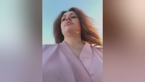 Media: A video of a young woman with long, wavy brown hair, wearing a light pink robe, captured from a low angle, with a blurred, gradient background transitioning from pale pink to light blue.