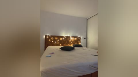 Media: A dimly lit, minimalist bedroom with a white wall, a wooden headboard adorned with fairy lights, a neatly made bed with two black pillows, and a closed closet door.