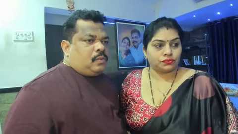 Media: Video of an Indian couple indoors, the man in a maroon shirt, the woman in a red and black saree, both with serious expressions, surrounded by framed family photos.